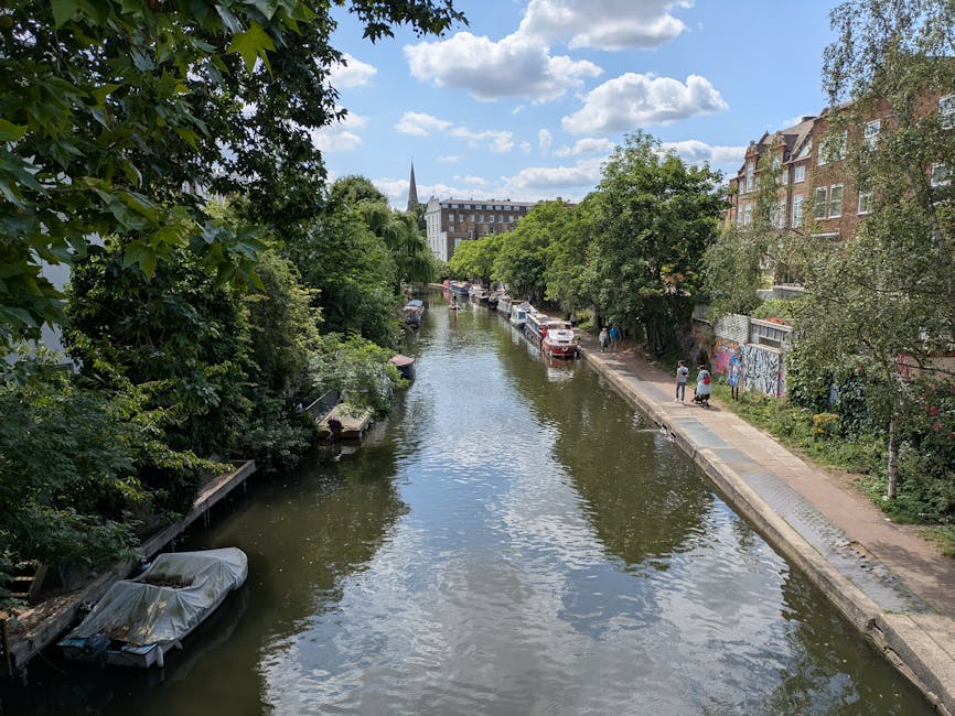 A narrow canal lined with lush green trees on both sides, with several boats moored along the edges, some covered with protective tarps. On the right side, there is a paved pedestrian path with a few individuals walking, and parked cars including a white van close to a building with graffiti-covered walls. The clear blue sky with scattered clouds is reflected in the calm water, and in the background, there are multi-storey brick buildings and a church steeple rising above the treetops. The scene captures a peaceful urban riverside, possibly in a residential area, suitable for house removals and furniture transport services as part of a home relocation process, supported by companies like Man and Van Pentonville.