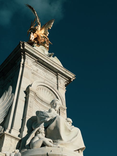 A detailed view of a historic marble monument located outdoors, featuring a large seated female sculpture with a child at her feet, both crafted from white marble with intricate carvings and lifelike facial features. The monument is set against a deep blue sky and topped with a gilded bronze sculpture of a winged figure holding a laurel wreath and a sword. The base of the monument includes decorative elements such as scrollwork and relief carvings. Surrounding the base are other smaller carved figures, some of which are partially visible. The scene appears illuminated by natural daylight, casting soft shadows on the sculptures. While not directly related to house removals, this image provides a static view of an outdoor monument that could be used to symbolize careful handling and transportation of valuable or delicate objects during a home relocation. For professional moving and furniture transport services, [COMPANY_NAME] offers expert assistance in packing and loading processes, ensuring safe transit of household items.