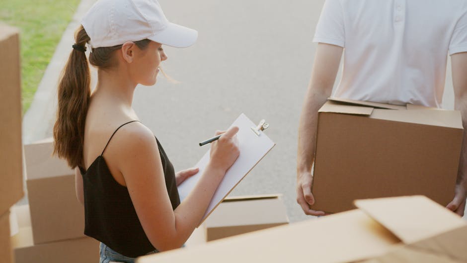 A young woman with long brown hair tied in a ponytail, wearing a white cap and a black strap top, is holding a clipboard and pen while standing outdoors near a moving van. She is observing a man, whos