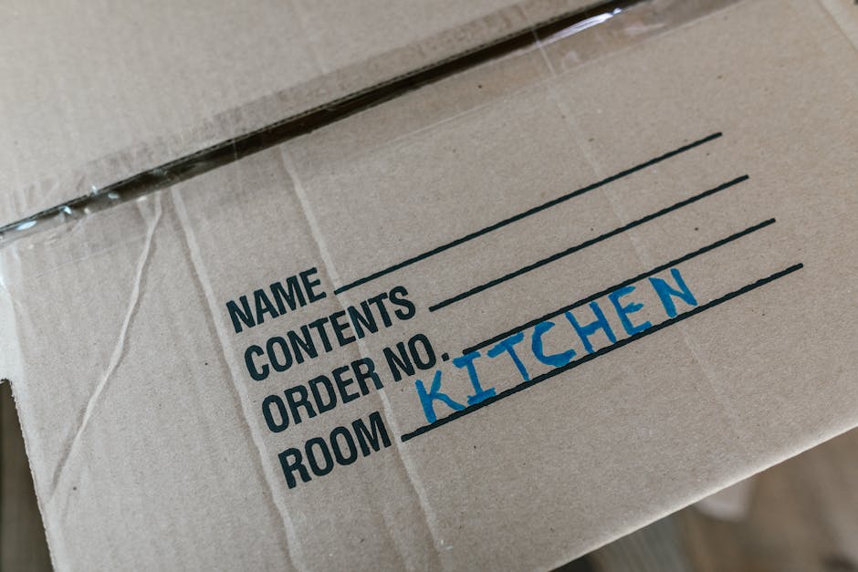 A woman smiling and holding a large cardboard box labeled 'GLASS' during a home relocation process. She is standing indoors in front of a textured light blue wall, surrounded by additional packing car