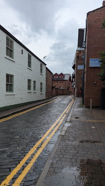 A narrow, cobbled street on a cloudy day, with a white painted building on the left featuring multiple windows, and a red brick building on the right with a blue sign. The street has double yellow lines along the edge and gently curves uphill. Paving slabs are visible on the sidewalk, and a small portion of a parked vehicle or moving van can be seen at the bottom right. The scene depicts an urban environment suitable for house removals or furniture transport, with no people present. During a home relocation, Man and Van Pentonville would carefully navigate this street for loading or unloading furniture, with packing materials, boxes, and possibly blankets required for safe transport of household items.