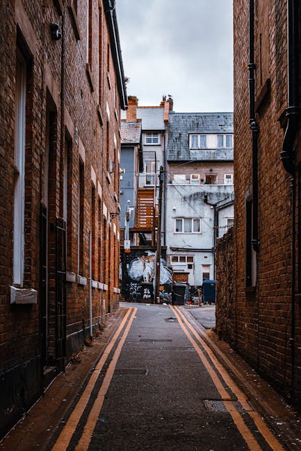 A narrow urban alleyway between two brick buildings, with a double yellow line marking the centre of the paved ground. At the end of the alley, there are residential buildings with a mix of brick and concrete facades, small windows, and a wooden external staircase leading up to an upper floor. The alley is slightly wet, possibly from recent rain, and features black drainpipes and utility boxes attached to the buildings' walls. In the background, a large black van is parked near the end of the alley, partially visible behind some stacked packaging materials and moving equipment, such as cardboard boxes wrapped in plastic, moving blankets, and straps. The scene suggests the site of a home relocation or furniture transport process, conducted by a professional removal service, with the alley serving as the access point for loading or unloading items during a house move.