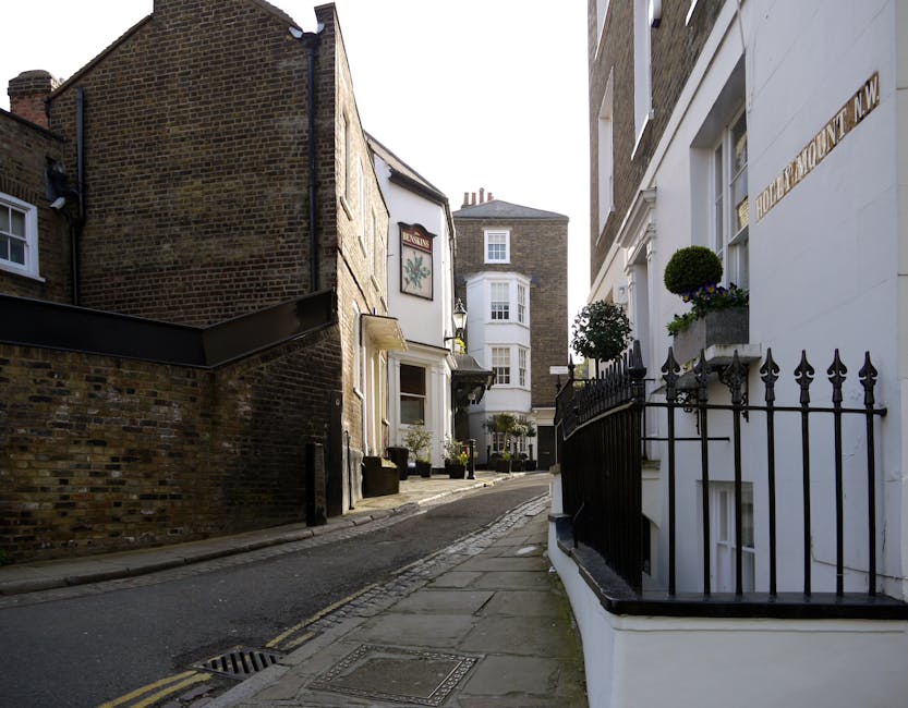 A narrow, cobbled street on a cloudy day, with a white painted building on the left featuring multiple windows, and a red brick building on the right with a blue sign. The street has double yellow lines along the edge and gently curves uphill. Paving slabs are visible on the sidewalk, and a small portion of a parked vehicle or moving van can be seen at the bottom right. The scene depicts an urban environment suitable for house removals or furniture transport, with no people present. During a home relocation, Man and Van Pentonville would carefully navigate this street for loading or unloading furniture, with packing materials, boxes, and possibly blankets required for safe transport of household items.